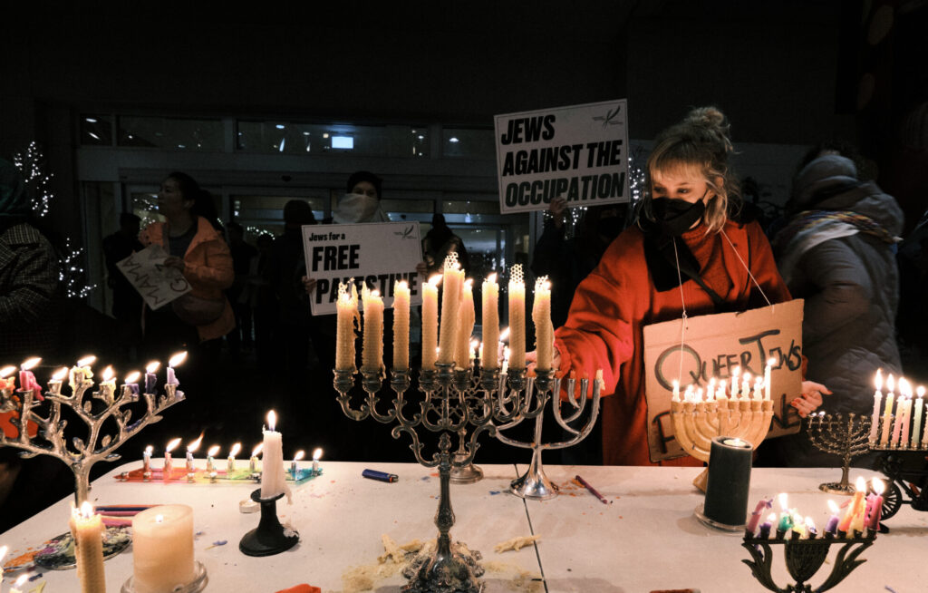 A Jewish person with an orange sweater, a black face mask and blonde shoulder length hair lights a menorah on a table full of lit menorahs, in front of a group of Jewish people holding signs.
