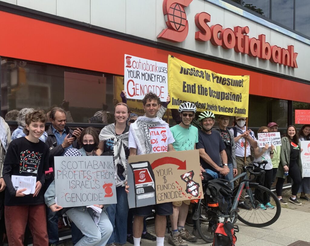 A group of IJV members and supporters stand in front of a Scotiabank branch holding signs calling out the bank's complicity in genocide.
