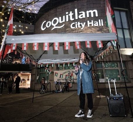 A Jewish woman with a blue coat and long curly hair makes a speech in front of Coquitlam City Hall.