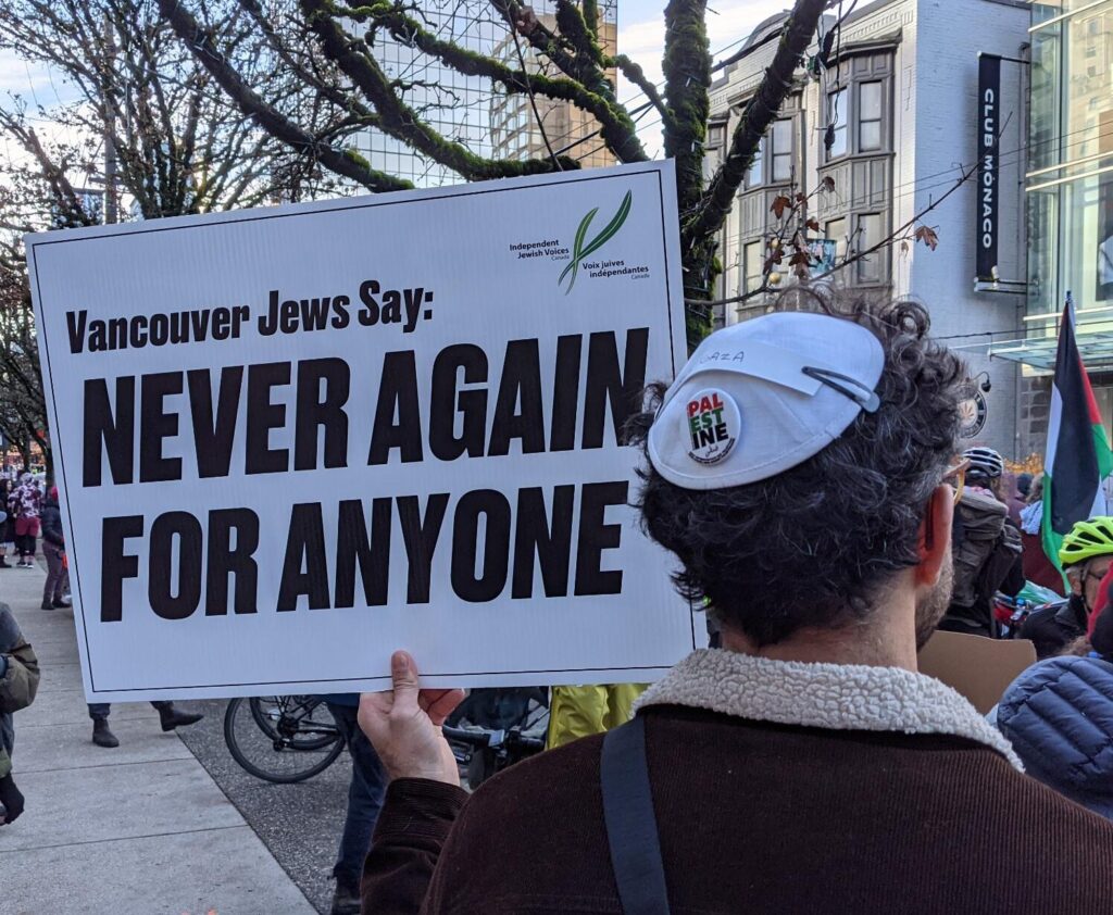 A man with curly brown hair and a white kippah with text "Free Gaza" and "Free Palestine" stands backwards and holds a sign that says "Vancouver Jews Say: Never Again for Anyone"